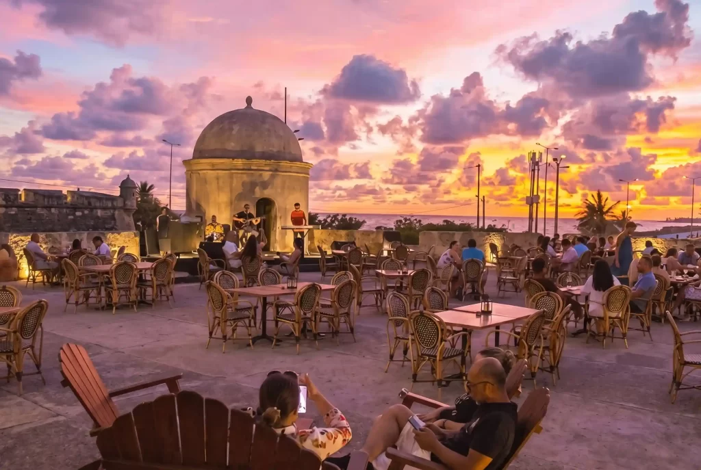 Rooftop bar on Cartagena's colonial walls at sunset with live music, illuminated watchtower and Caribbean Sea view.