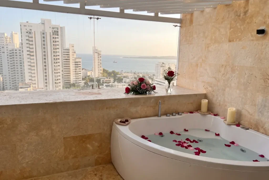 Jacuzzi decorado con pétalos de rosa y velas junto a un ventanal con vista panorámica a los edificios y al mar en Cartagena.